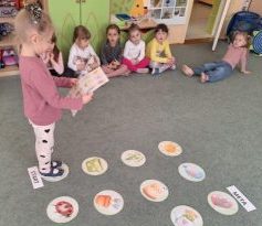 Na dywanie rozłożone są wiosenne emblematy. Dziecko porusza się po nich zgodnie z obrazkową instrukcją trzymaną w rękach.