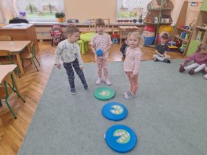Troje dzieci stoi wokół kolorowych podkładek na których ułożone są liście i kwiaty lilii wodnych. Jedno dziecko trzyma dużą kostkę.