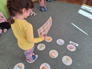 Na dywanie rozłożone są wiosenne emblematy. Dziecko porusza się po nich zgodnie z obrazkową instrukcją trzymaną w rękach.