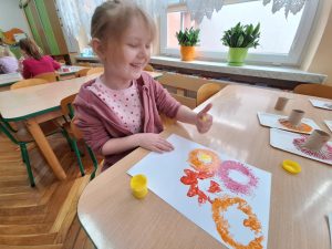 Dziewczynka przy stoliku. Wykonują prace plasytyczną - odbijają nacięte rolki po papierze tworząc wiosenne kwiaty.