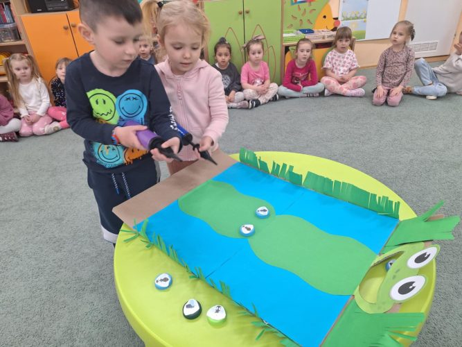 Dwójka dzieci trzyma pompki do balonów. Przesuwają muchy zrobione z zakrętek, po kolorowej trasie. Karmią żabkę.