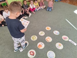 Na dywanie rozłożone są wiosenne emblematy. Dziecko porusza się po nich zgodnie z obrazkową instrukcją trzymaną w rękach.