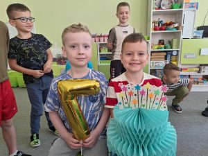Solenizanci siedzą na krzesełkach, jeden z nich trzyma balon w kształcie cyfry 7, a drugi papierowy tort.