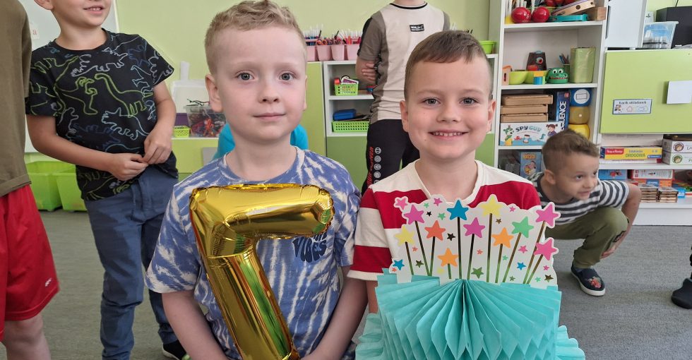 Solenizanci siedzą na krzesełkach, jeden z nich trzyma balon w kształcie cyfry 7, a drugi papierowy tort.