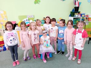 Solenizantka siedzi na krześle. Obok niej stoją wszystkie koleżanki z grupy.