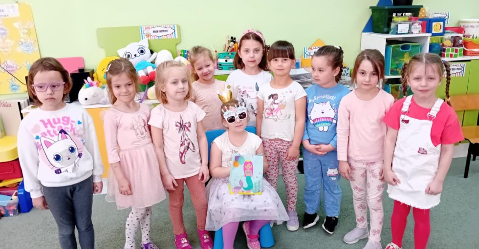 Solenizantka siedzi na krześle. Obok niej stoją wszystkie koleżanki z grupy.