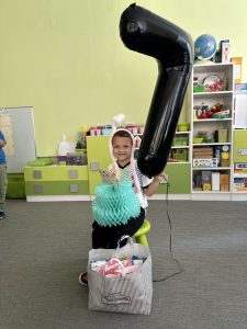 Chłopiec siedzi na krześle. na głowie ma opaskę z króliczymi uszami. W lewej ręce trzyma duży balon w kształcie cyfry 7. Na kolanach trzyma papierowy tort. Przed nim stoi torba ze słodkościami.