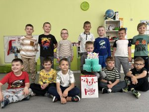 Solenizant siedzi na krześle. wokół niego stoją i siedzą koledzy z grupy.