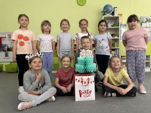 Solenizant siedzi na krześle. wokół niego stoją i siedzą koleżanki z grupy.