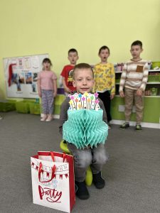 Solenizant siedzi na krześle, trzyma papierowy tort, w tle stoją dzieci.