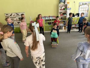 Chłopiec siedzi na krześle. Trzyma papierowy tort. Wokół  otacza go grupa dzieci.