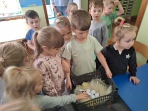 Chłopiec trzyma skrzynkę z plastikowymi jajkami. Dziewczynka wyjmuje ze skrzynki kartkę z zagadką. Reszta dzieci stoi wokół.