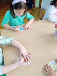 Dziewczynka siedzi przy stoliku i koloruje obrazek przedstawiający kosz na plastiki.