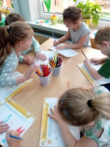 Dzieci siedzą przy stolikach i łączą z plastikową butelką inne plastikowe przedmioty.
