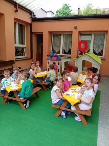 Dzieci siedzą przy stolikach na tarasie i jedzą podwieczorek.
