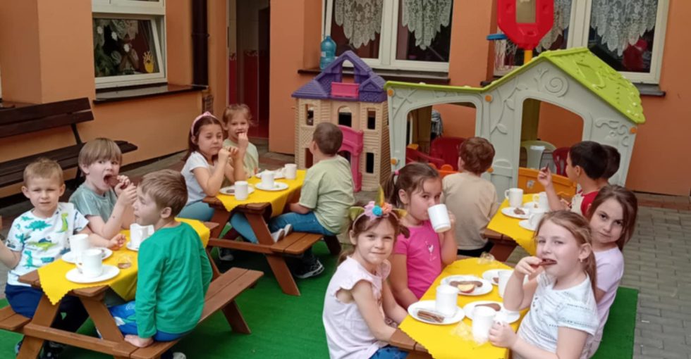 Dzieci siedzą przy stolikach na tarasie i jedzą podwieczorek.