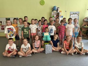 Solenizant siedzi na środku, w rękach trzyma papierowy tort. wokół niego stoją koleżanki i koledzy  z grupy.