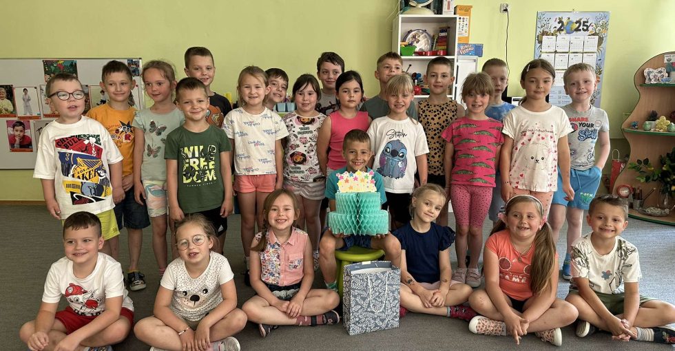 Solenizant siedzi na środku, w rękach trzyma papierowy tort. wokół niego stoją koleżanki i koledzy z grupy.