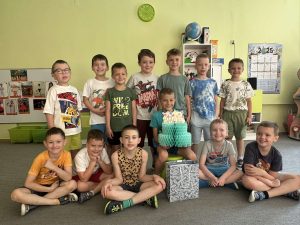Solenizant siedzi na środku, w rękach trzyma papierowy tort. wokół niego stoją koledzy  z grupy.