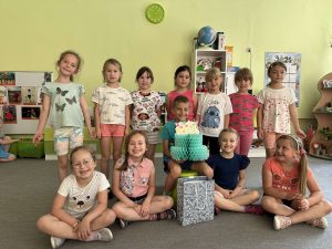 Solenizant siedzi na środku, w rękach trzyma papierowy tort. wokół niego stoją koleżanki z grupy.