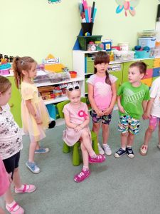 Dzieci stoją w kole i śpiewają siedzącej na krześle Solenizantce Sto lat.
